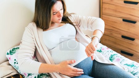 Téléchargez les photos : Contractions douloureuses gravides. Femme enceinte regardant l'horloge, tenant le ventre de bébé. Temps d'accouchement, douleurs de contractions. Grossesse, médecine concept de soins de santé - en image libre de droit