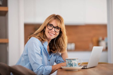 Photo for Close-up of middle aged woman sitting at home and using earphone and laptop while having video call. Home office. - Royalty Free Image