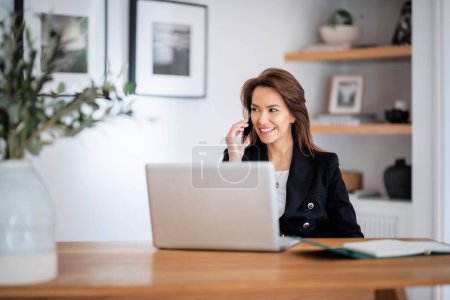 Photo for Shot of an attractive businesswoman using a laptop and having a call while working in her home office. Confident female wearing blazer. - Royalty Free Image