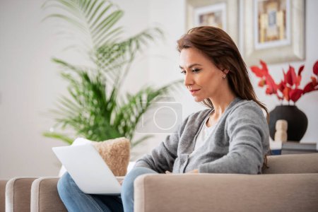 Photo for Shot of an attractive middle aged woman sitting on armchair while using laptop. Beautiful female wearing casual clothes. - Royalty Free Image