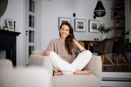 Foto de Retrato de una hermosa mujer de pelo largo con ropa casual mientras se relaja en un sillón en casa. - Imagen libre de derechos