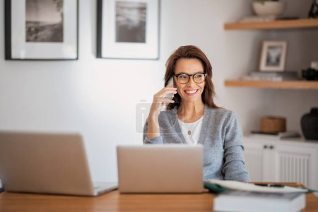 Photo for Attractive woman wearing eyewear and casual clothes while working from home. Business woman using laptops and making a call. Home office. - Royalty Free Image