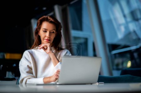 Téléchargez les photos : Femme d'affaires d'âge moyen utilisant un ordinateur portable tout en étant assis au bureau au bureau. Femme d'affaires exécutive portant un pull. - en image libre de droit