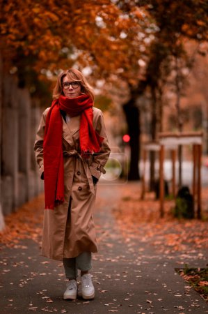Photo for Fashionable middle aged woman walking in the street and enjoying autumn day. Attractive female wearing scarf and trench coat outdoor. - Royalty Free Image