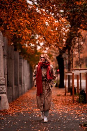Foto de Mujer de mediana edad de moda caminando por la calle y disfrutando del día de otoño. Atractiva mujer con bufanda y gabardina al aire libre. - Imagen libre de derechos