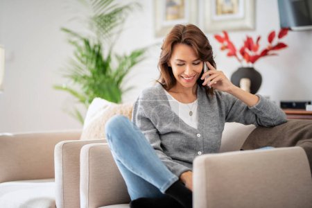 Photo for Portrait of beautiful long haired woman relaxing in an armchair and using mobile phone. Smiling female wearing casual clothes at home and having a call. - Royalty Free Image