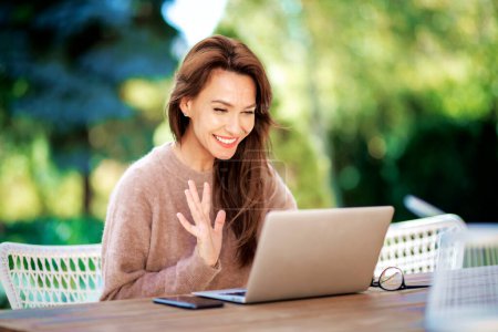 Téléchargez les photos : Femme d'âge moyen confiante avec un ordinateur portable assis sur le balcon et utilisant un ordinateur portable. Attrayant femelle agitant sa main lors d'un appel vidéo. - en image libre de droit
