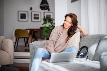 Photo for Shot of an attractive middle aged woman sitting on sofa and using laptop. Beautiful female wearing casual clothes. - Royalty Free Image
