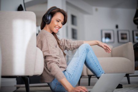 Photo for Close-up of beautiful middle aged woman with headphones using a laptop while having video call. Attractive female wearing sweater and blue jeans. - Royalty Free Image