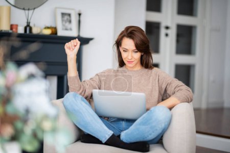 Photo for Shot of an attractive middle aged woman sitting on armchair while using laptop. Beautiful female wearing casual clothes. - Royalty Free Image