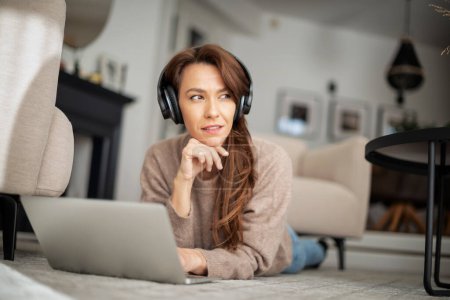Téléchargez les photos : Pleine longueur de belle femme d'âge moyen avec écouteurs utilisant un ordinateur portable tout en naviguant sur Internet à la maison. - en image libre de droit