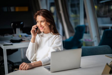 Photo for Middle aged business woman making a call and using laptop while sitting at desk at the office. Executive businesswoman wearing sweater. - Royalty Free Image