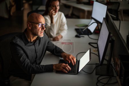 Photo for Shot of a businessman looking thoughtfully while sitting at his desk in the office in late evening. Confident businesswoman sitting next to him and working together. Teamwork. - Royalty Free Image
