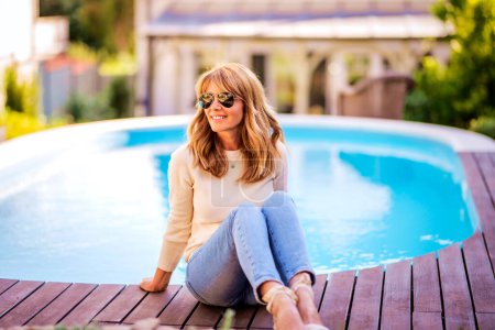 Photo for Full length of middle aged woman wearing sunglasses and smiling while relaxing by the poolside at home. - Royalty Free Image