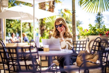 Photo for Shot of happy middle aged woman having a call while sitting at outdoor cafe. Confident businessman wearing casual clothes and using a laptop while working online. - Royalty Free Image