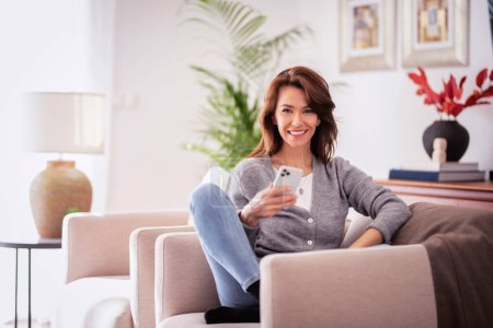 Photo for Attractive brunette haired woman wearing casual clothes while relaxing at home and using a smartphone. Smiling female text messaging. - Royalty Free Image