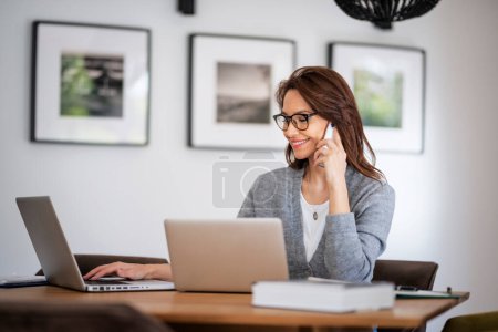 Foto de Mujer atractiva que usa gafas y ropa casual mientras trabaja desde casa. Mujer de negocios usando computadoras portátiles y haciendo una llamada. Oficina en casa. - Imagen libre de derechos