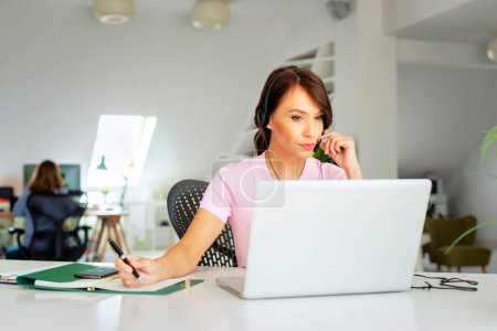 Foto de Asistente de servicio al cliente que usa auriculares mientras está sentada detrás de su computadora portátil y trabaja en el centro de llamadas. - Imagen libre de derechos