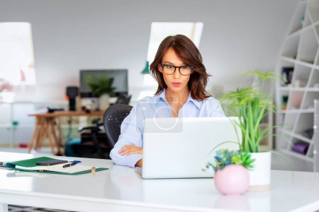Photo for Confident middle aged businesswoman sitting at office desk behind her laptop and having video call. - Royalty Free Image
