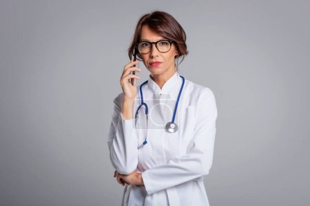 Photo for A mid aged female doctor wearing a white lab coat and using mobile phone while standing at isolated background. Copy space. - Royalty Free Image