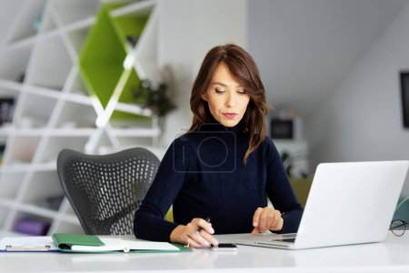 Téléchargez les photos : Jolie femme d'affaires d'âge moyen utilisant un ordinateur portable et un téléphone portable tout en travaillant au bureau. Femme d'affaires exécutive portant un pull. - en image libre de droit
