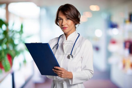 Photo for A mid aged female doctor wearing a white lab coat while standing inside a hospital building. She is looking at the camera and smiling. Attractive female doctor holding clipboard in her hand while standing at the clinic. - Royalty Free Image
