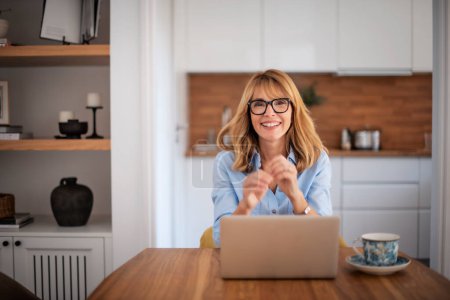Photo for Happy mid aged woman sitting at table and using laptop while working from home. Home office. - Royalty Free Image