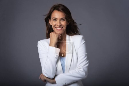 Photo for Close-up of an attractive middle aged woman with toothy smile wearing blazer while sitting at isolated dark background. Copy space. Studio shot. - Royalty Free Image