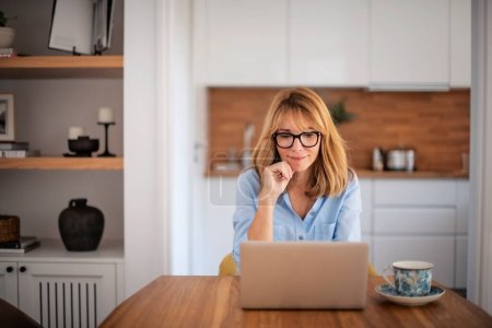 Foto de Hermosa mujer de mediana edad utilizando el ordenador portátil mientras trabaja desde casa. Oficina en casa. - Imagen libre de derechos