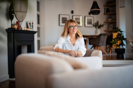 Foto de Retrato de una atractiva mujer de mediana edad que se relaja en un sillón en casa. Mujer de pelo rubio con gafas y camisa blanca. - Imagen libre de derechos