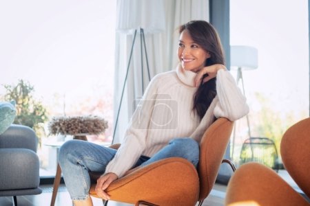 Photo for An attractive brunette haired woman relaxing in an amrchair at home. Beautiful female wearing sweater and blue jeans. - Royalty Free Image