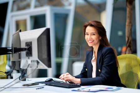 Photo for Middle aged business woman using computer while sitting at desk at the office. Professional female working on financial statements. - Royalty Free Image