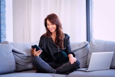 Foto de Attractive long haired woman wearing black shirt and jeans while sitting at home in an armchair. Smiling female text messaging. - Imagen libre de derechos