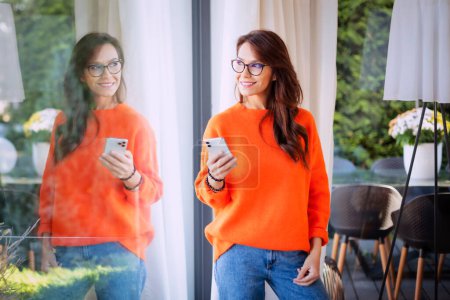 Téléchargez les photos : Attractive long haired woman wearing orange sweater and blue jeans while standing at the window and looking out. Cheerful smiling female text messaging. - en image libre de droit