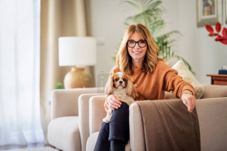 Foto de Portrait of attractive middle aged woman relaxing in an armchair at home with her king charles cavalier puppy. - Imagen libre de derechos
