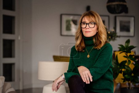 Foto de Portrait of attractive middle aged woman daydreaming in an armchair at home. Blond haired female wearing eyeglasses and green turtleneck sweater. - Imagen libre de derechos