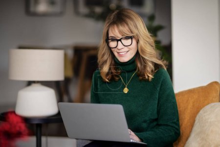 Photo for Portrait of attractive middle aged woman sitting at home and using laptop. Blond haired female wearing eyeglasses and turtleneck sweater. Home office. - Royalty Free Image