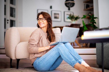Photo for Close-up of an attractive middle aged woman sitting on floor while using laptop. Beautiful female wearing casual clothes. - Royalty Free Image