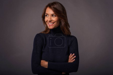Photo for Close-up of an attractive middle aged woman with toothy smile wearing black turtleneck sweater while standing at isolated dark background. Copy space. Studio shot. - Royalty Free Image