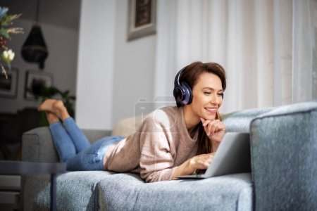 Foto de Hermosa mujer de mediana edad con auriculares utilizando un ordenador portátil mientras navega por Internet. Mujer sonriente relajándose en el sofá en casa. - Imagen libre de derechos