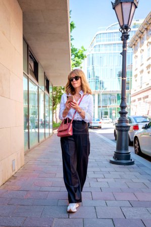 Téléchargez les photos : Full length of an attractive woman wearing sunglasses and text messaging while walking on sidewalk in the city. - en image libre de droit