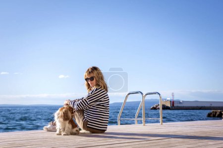 Foto de Vista trasera de una mujer con suéter a rayas y gafas de sol mientras está sentada con su lindo perro en el embarcadero junto al mar. - Imagen libre de derechos