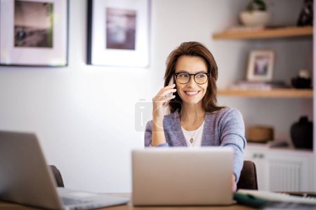Photo for Happy woman using mobile phone and laptops for work. Confident business woman working from home. Home office. - Royalty Free Image
