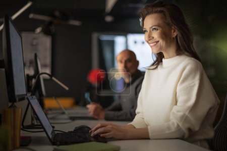 Téléchargez les photos : Photo d'une femme d'affaires souriante assise à table avec un ordinateur portable au bureau en fin de soirée. Sa collègue assise à côté d'elle. - en image libre de droit
