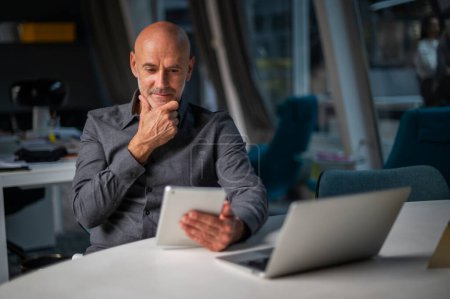 Téléchargez les photos : Prise de vue d'un homme d'affaires réfléchi utilisant une table numérique et un ordinateur portable assis au bureau et travaillant dans un bureau moderne. - en image libre de droit