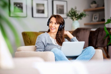 Foto de Beautiful middle aged woman with headphone using a laptop while listening music. Attractive caucasian female relaxing in an armchair at home. - Imagen libre de derechos