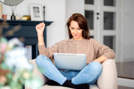 Foto de An attractive middle aged woman sitting in armchair at home while using laptop. Beautiful female wearing casual clothes. Home office. - Imagen libre de derechos