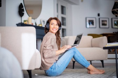 Photo for Beautiful middle aged woman with headphone using a laptop while listening music. Attractive caucasian female wearing casual clothes and relaxing on the floor at home. - Royalty Free Image