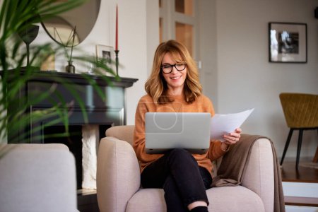 Foto de Middle aged woman using laptop and holding document in her hand while sitting at home and working. Home office. - Imagen libre de derechos