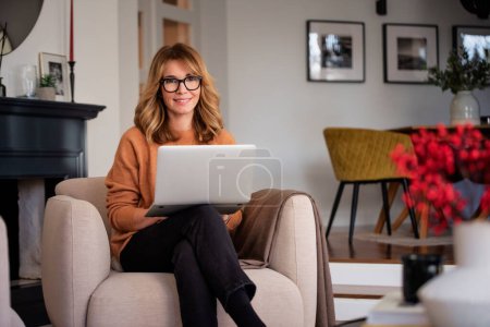 Foto de An attractive middle aged woman sitting in the armchair at home and using laptop. Beautiful female wearing casual clothes while working from home. Home office. - Imagen libre de derechos